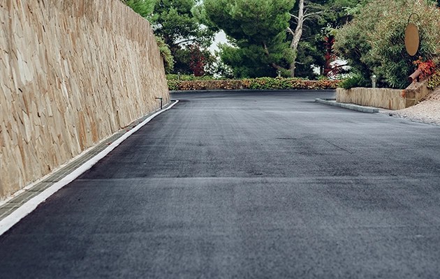 KSW A newly paved asphalt road curves gently to the left, bordered by a stone wall on the left and greenery on the right. Denver Metro