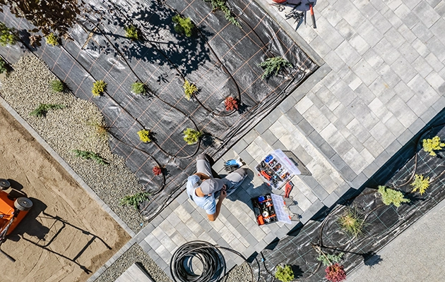 KSW A person is sitting on the ground next to gardening tools and small plants arranged on a weed barrier in a paved and gravel area. Denver Metro