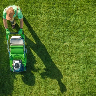 KSW A person wearing a hat, gloves, and green outfit is mowing a neatly trimmed lawn with a green lawnmower on a sunny day. Denver Metro