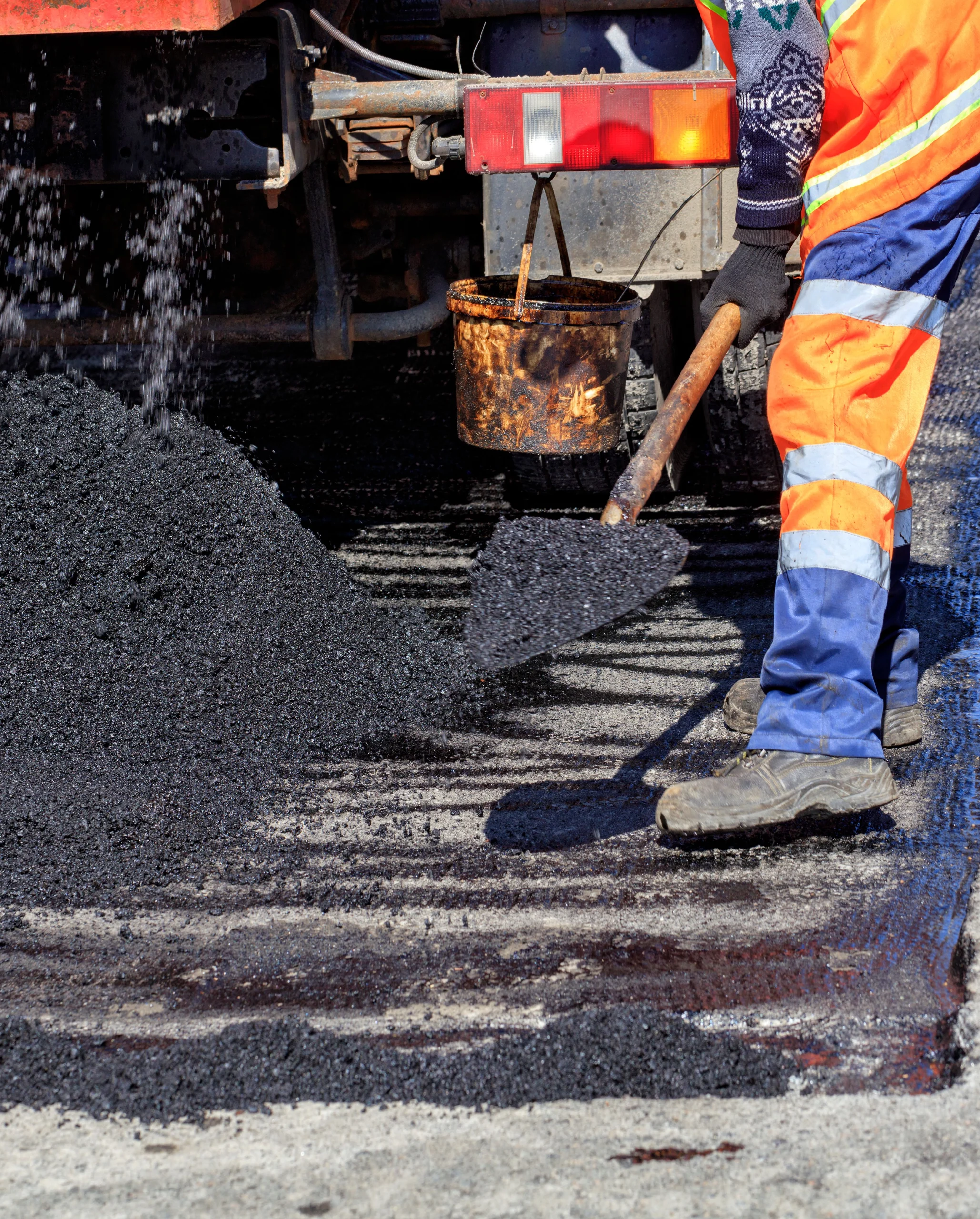 KSW Worker in orange and blue uniform shoveling hot asphalt from a truck onto a road surface for repairs. Denver Metro